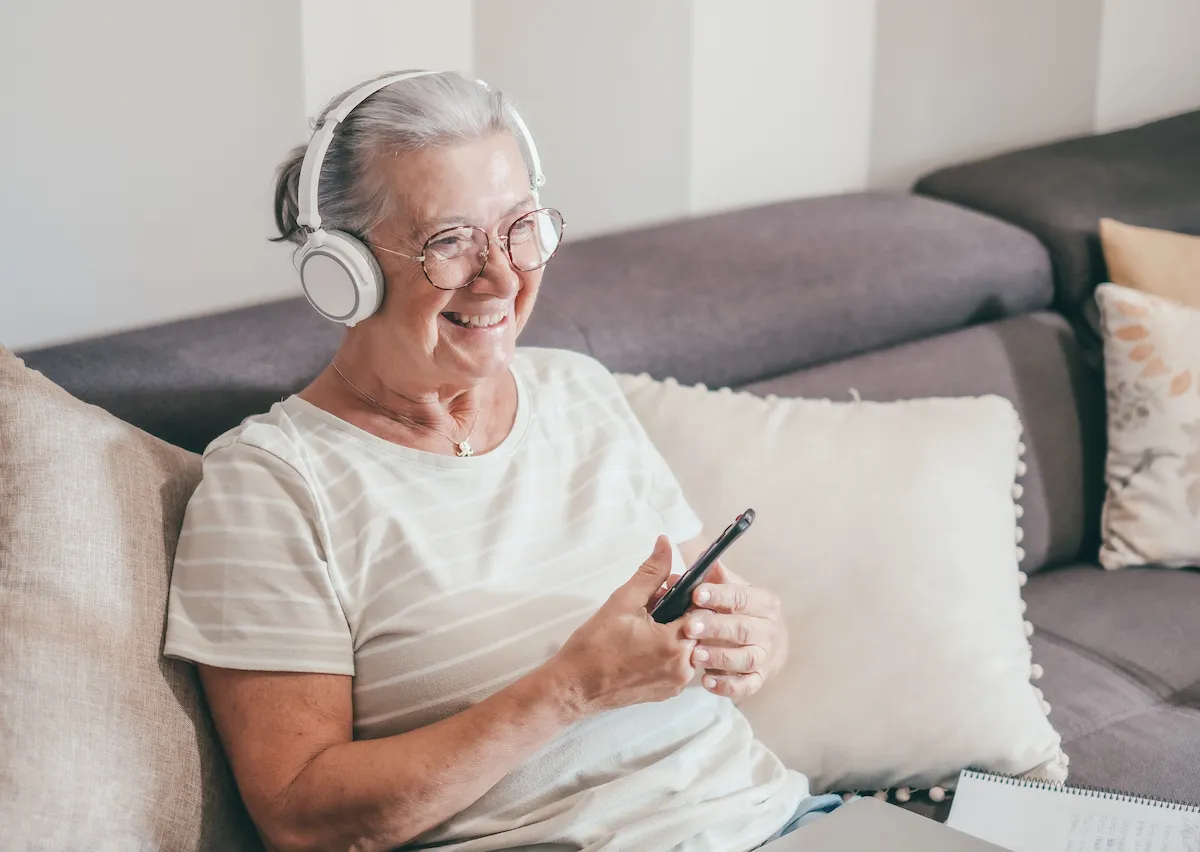 Mulher idosa sentada em um sofá com fones de ouvido e telefone, sorrindo em um ambiente confortável e acolhedor.