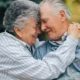 Casal de idosos abraçados ao ar livre, sorrindo, com um tablet para exercícios de reabilitação cognitiva no Dia dos Namorados.