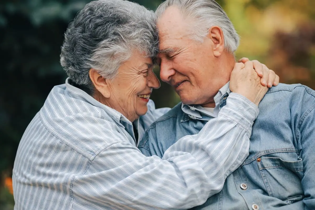 Casal de idosos abraçados ao ar livre, sorrindo, com um tablet para exercícios de reabilitação cognitiva no Dia dos Namorados.