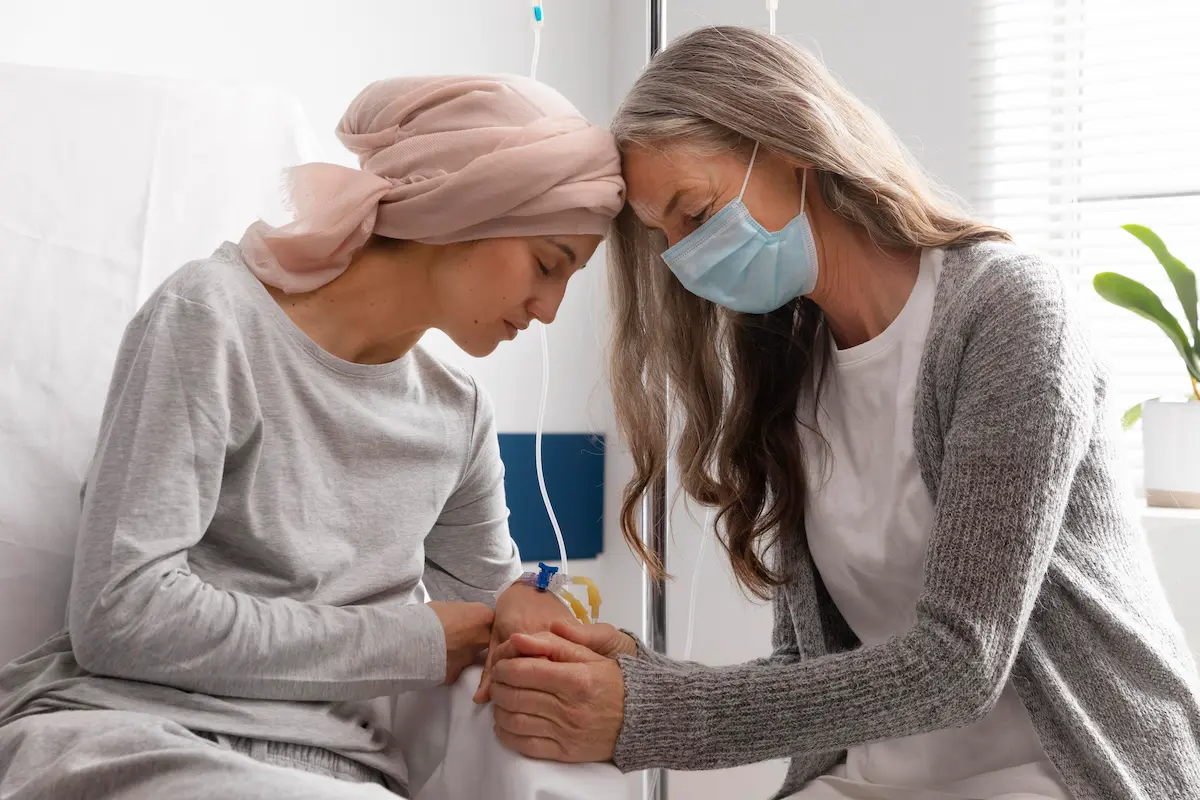 Duas mulheres em um hospital: uma com lenço na cabeça e a outra com máscara, demonstram apoio emocional.