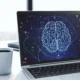 O laptop aberto mostra um cérebro de design gráfico feito com circuitos; ao lado, uma xícara, livros e luz natural.