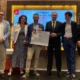 Grupo de pessoas posando em um evento formal; no centro, alguém segura um certificado, em frente a um cartaz vermelho.