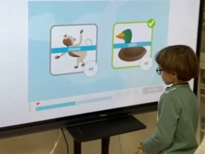 Uma criança interage em frente a uma tela durante uma sessão do NeuronUP; vê-se um macaco e um pato, com um exercício de palavras que começam com PA.