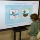 Uma criança interage em frente a uma tela durante uma sessão do NeuronUP; vê-se um macaco e um pato, com um exercício de palavras que começam com PA.