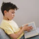 Criança sentada no sofá, camiseta amarela e cabelo castanho bagunçado, lendo um livro; ambiente moderno e tranquilo.