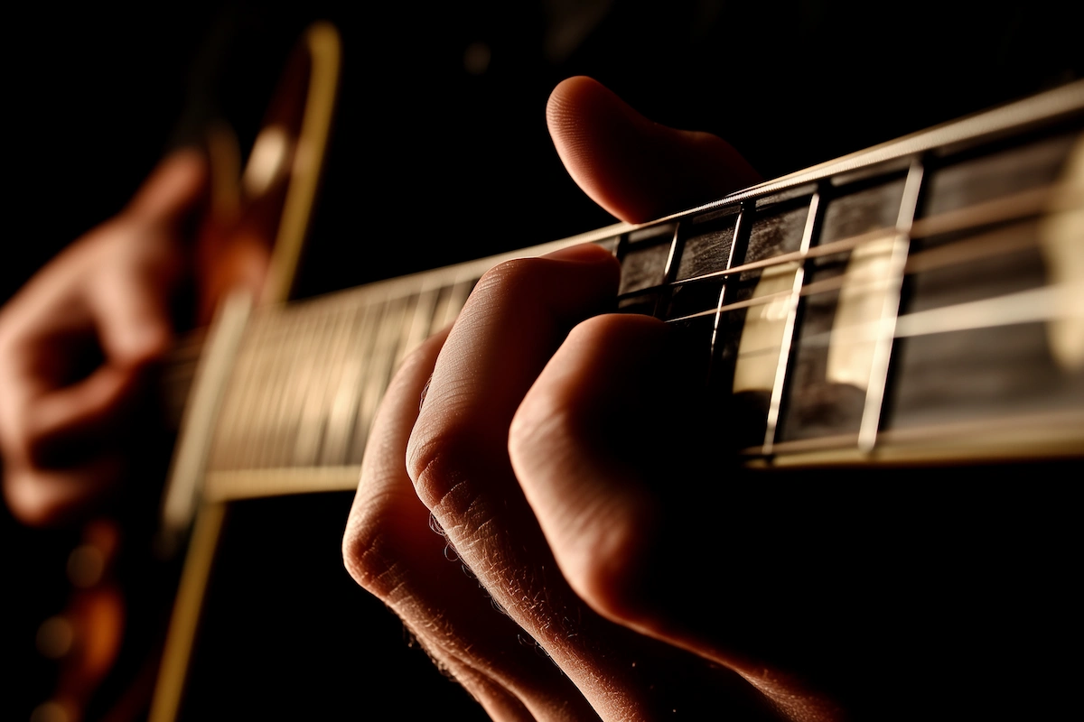 Mão pressionando as cordas no braço de uma guitarra, com foco nos dedos e nos trastes, em um fundo escuro.