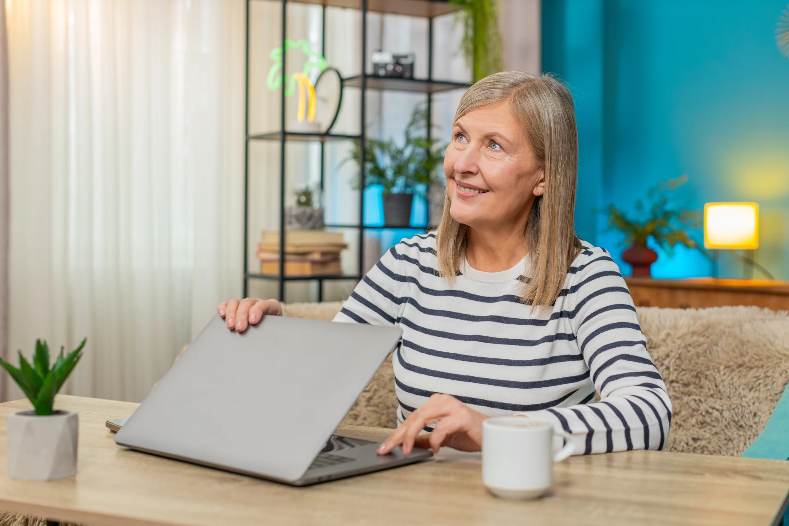 Mulher adulta em frente a um notebook sobre uma mesa, com uma xícara e uma planta, em um ambiente iluminado; teleterapia de linguagem para afasia.