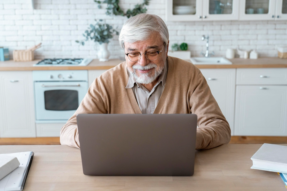 Homem idoso sentado diante de um notebook em uma cozinha moderna, sorrindo; fogão, tijolos brancos e plantas ao fundo.