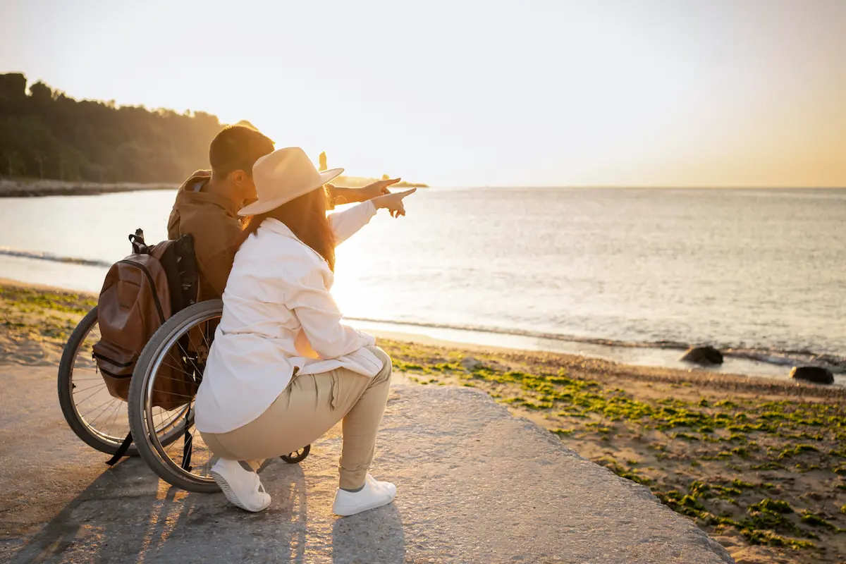 Paar am Strand: Eine Person im Rollstuhl stützt sich auf den Arm der anderen; beide blicken bei Sonnenuntergang auf das Meer, umgeben von Vegetation.