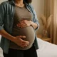 Schwangere Frau im Profil, die in einem warmen Innenraum steht, eine Hand auf dem Bauch und sanftes natürliches Licht.