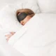 Person im Bett mit schwarzer Schlafmaske und weißer Decke, eine ruhige Szene zur Darstellung von Schlaf und kognitiver Wachheit.