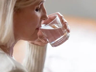 Blonde Frau hält ein Glas Wasser an die Lippen und trinkt in einer sanft beleuchteten Umgebung mit hellem Pullover.