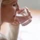 Blonde Frau hält ein Glas Wasser an die Lippen und trinkt in einer sanft beleuchteten Umgebung mit hellem Pullover.