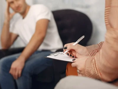 Erwachsener Mann in einer klinischen Sprechstunde; Nahaufnahme einer Hand, die in ein Notizbuch schreibt, während emotionale Intelligenz bei Schizophrenie thematisiert wird.