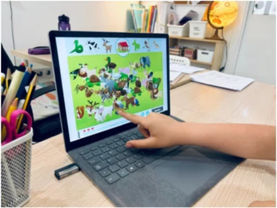 Tablette mit farbenfroher NeuronUP-Oberfläche und Tieren, die Hand eines Kindes zeigt auf den Bildschirm, Bleistifte und ein Luftballon auf dem Tisch.