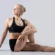 Frau in sitzender Yoga-Haltung, ein Bein seitlich ausgestreckt, das andere angewinkelt, mit zusammengebundenem Haar und ärmellosem T-Shirt.