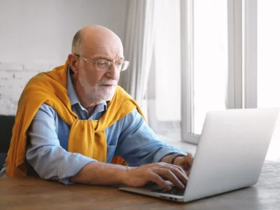 Älterer Mann mit grauem Haar und Bart sitzt in einer modernen Klinik vor einem Laptop und arbeitet konzentriert mit NeuronUP.