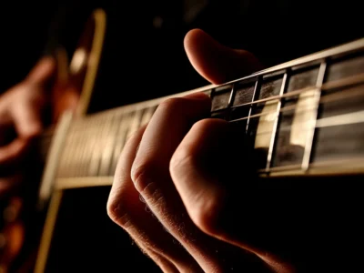 Hand, die Saiten am Hals einer Gitarre drückt, mit Fokus auf Finger und Bünde vor dunklem Hintergrund.