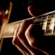 Hand, die Saiten am Hals einer Gitarre drückt, mit Fokus auf Finger und Bünde vor dunklem Hintergrund.