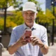 Lächelnder älterer Mann hält ein Mobiltelefon im Vordergrund; kariertes Hemd, weiße Mütze und sonniger urbaner Hintergrund.
