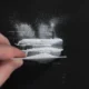 Dunkle Oberfläche mit verstreutem weißem Pulver; eine Hand glättet das Pulver mit einer Karte und erzeugt Muster auf dem Hintergrund.