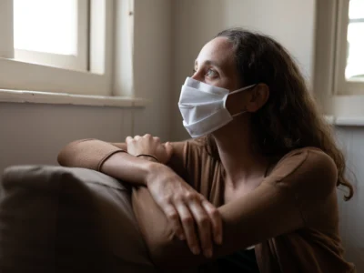 Frau mit weißer Maske sitzt auf einem Sofa und blickt bei sanfter Beleuchtung in einem schlichten Innenraum aus dem Fenster.
