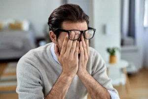 Mann mit Brille und hellem Pullover bedeckt in häuslicher Umgebung sein Gesicht mit den Händen und wirkt gestresst.