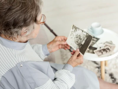 Ältere Frau betrachtet im Sitzen ein Schwarz-Weiß-Foto; sie trägt eine Brille, hat graues Haar und einen Gehstock; auf dem Tisch liegen weitere Fotos und eine Tasse.