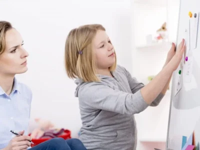 Mädchen interagiert mit einer Tafel, während eine Therapeutin sie in einer pädagogischen Umgebung beobachtet, die die soziale Perspektive veranschaulicht.