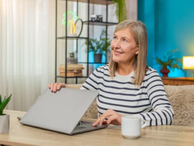 Erwachsene Frau sitzt in heller Umgebung mit Tasse und Pflanze an einem Laptop; Sprachtherapie bei Aphasie per Teletherapie.