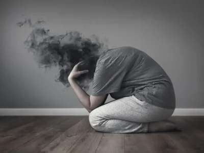 Lehrreiche Darstellung einer verzweifelten, knienden Person mit verschwommenem Kopf in einer dunklen Wolke; grauer Hintergrund und Holzoberfläche.