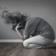 Lehrreiche Darstellung einer verzweifelten, knienden Person mit verschwommenem Kopf in einer dunklen Wolke; grauer Hintergrund und Holzoberfläche.