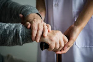 Das Bild zeigt Hände: Eine ältere Person mit Gehstock erhält Unterstützung von einer Betreuungsperson vor Bildschirmen mit NeuronUP; es veranschaulicht Fürsorge und Zusammenarbeit bei der kognitiven Stimulation.