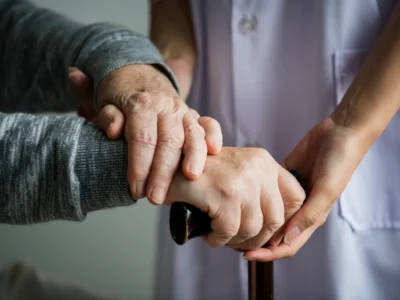 Das Bild zeigt Hände: Eine ältere Person mit Gehstock erhält Unterstützung von einer Betreuungsperson vor Bildschirmen mit NeuronUP; es veranschaulicht Fürsorge und Zusammenarbeit bei der kognitiven Stimulation.