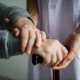 Das Bild zeigt Hände: Eine ältere Person mit Gehstock erhält Unterstützung von einer Betreuungsperson vor Bildschirmen mit NeuronUP; es veranschaulicht Fürsorge und Zusammenarbeit bei der kognitiven Stimulation.