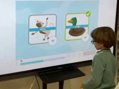 Kind interagiert während einer NeuronUP-Sitzung mit einem Bildschirm; ein Affe und eine Ente sowie eine Übung zu Wörtern mit PA-Anfang sind zu sehen.