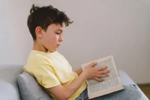 Kind mit gelbem T-Shirt und zerzaustem braunem Haar sitzt auf einem Sofa und liest ein Buch in einer modernen, ruhigen Umgebung.