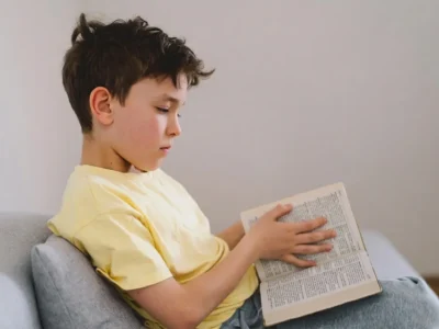 Kind mit gelbem T-Shirt und zerzaustem braunem Haar sitzt auf einem Sofa und liest ein Buch in einer modernen, ruhigen Umgebung.