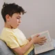 Kind mit gelbem T-Shirt und zerzaustem braunem Haar sitzt auf einem Sofa und liest ein Buch in einer modernen, ruhigen Umgebung.