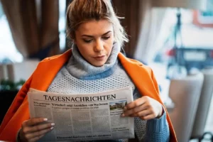 Frau mit orangefarbenem Mantel liest in einer warmen Umgebung bei sanfter Beleuchtung eine Zeitung.