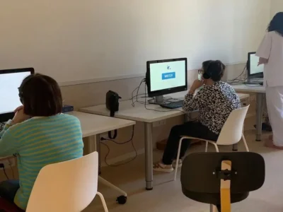 Raum mit mehreren FEM-Nutzerinnen und -Nutzern an Computern, die NeuronUP verwenden und Kopfhörer tragen; eine stehende Person beaufsichtigt sie.