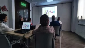 Gruppe von Teilnehmenden in einem Schulungsraum betrachtet eine Projektion mit farbigen Kreisen und Zahlen; auf dem Tisch steht ein Laptop, durch die Fenster fällt Tageslicht.