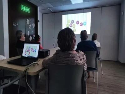 Gruppe von Teilnehmenden in einem Schulungsraum betrachtet eine Projektion mit farbigen Kreisen und Zahlen; auf dem Tisch steht ein Laptop, durch die Fenster fällt Tageslicht.