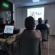 Gruppe von Teilnehmenden in einem Schulungsraum betrachtet eine Projektion mit farbigen Kreisen und Zahlen; auf dem Tisch steht ein Laptop, durch die Fenster fällt Tageslicht.