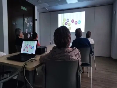 Gruppe von Teilnehmenden in einem Schulungsraum betrachtet eine Projektion mit farbigen Kreisen und Zahlen; auf dem Tisch steht ein Laptop, durch die Fenster fällt Tageslicht.