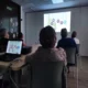 Gruppe von Teilnehmenden in einem Schulungsraum betrachtet eine Projektion mit farbigen Kreisen und Zahlen; auf dem Tisch steht ein Laptop, durch die Fenster fällt Tageslicht.