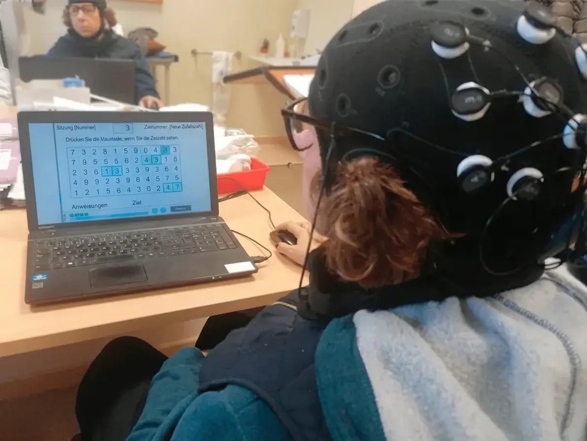 Person mit EEG-Kappe vor einem Laptop; der Bildschirm zeigt während einer tDCS-Sitzung mit NeuronUP ein Zahlenraster.