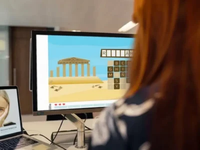 Person mit rötlichem Haar vor einem Bildschirm mit einem Wort- und Buchstabenspiel und einem Symbol eines griechischen Tempels in einem Büro.
