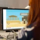 Person mit rötlichem Haar vor einem Bildschirm mit einem Wort- und Buchstabenspiel und einem Symbol eines griechischen Tempels in einem Büro.