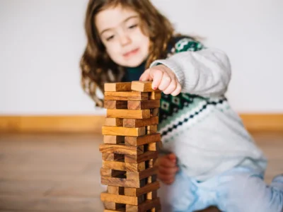 Kleines Mädchen spielt mit einem Turm aus Holzklötzen und legt einen Klotz auf die Spitze, neutraler Hintergrund.